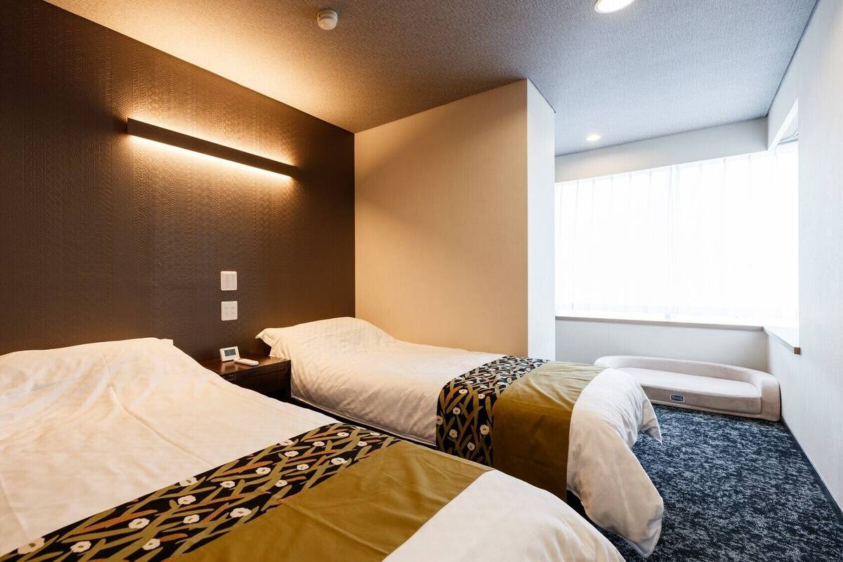 Habitación de hotel con dos camas, una mesita de noche con lámpara, una ventana con persianas y suelo alfombrado.