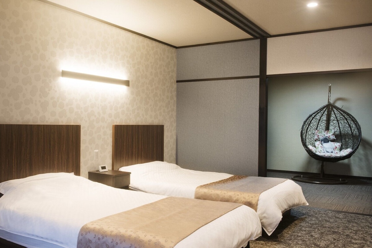 Hotellihuone, jossa on kaksi sänkyä, riipputuoli ja seinään kiinnitetty valaisin.