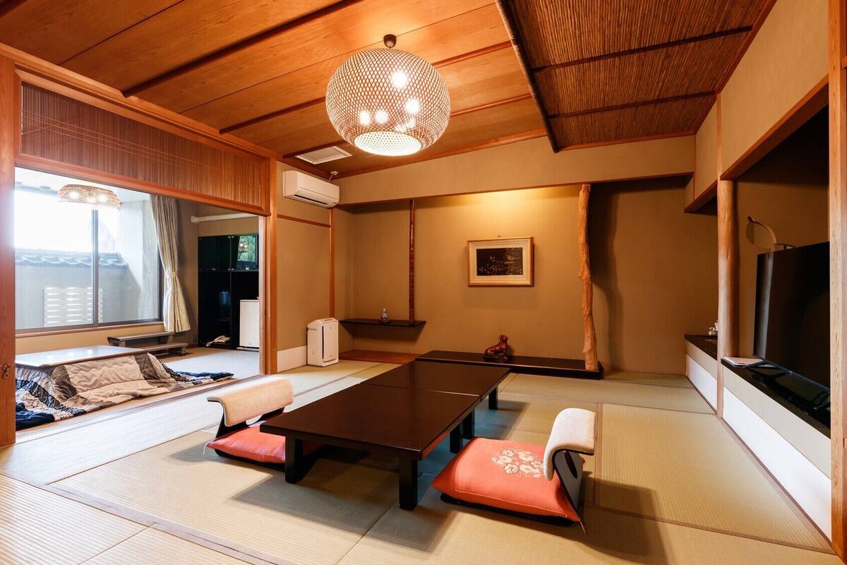 Una habitación tradicional japonesa con suelo de tatami, una mesa baja con cojines, una lámpara colgante y un televisor a la derecha.
