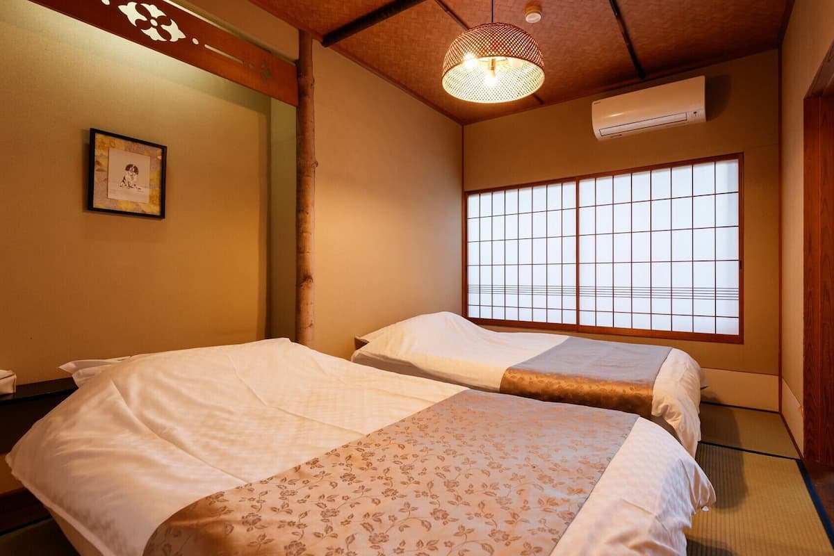 Perinteinen japanilaistyylinen huone, jossa on tatamimatto, kaksi kukka-kuvioisilla tekstiileillä varustettua sänkyä, shoji-tyylinen ikkuna ja riippuvalaisin.