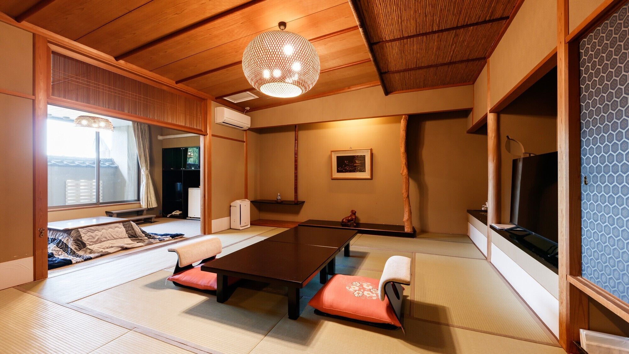 A traditional Japanese room with tatami flooring, a low table with cushions, a hanging light fixture, and a television.