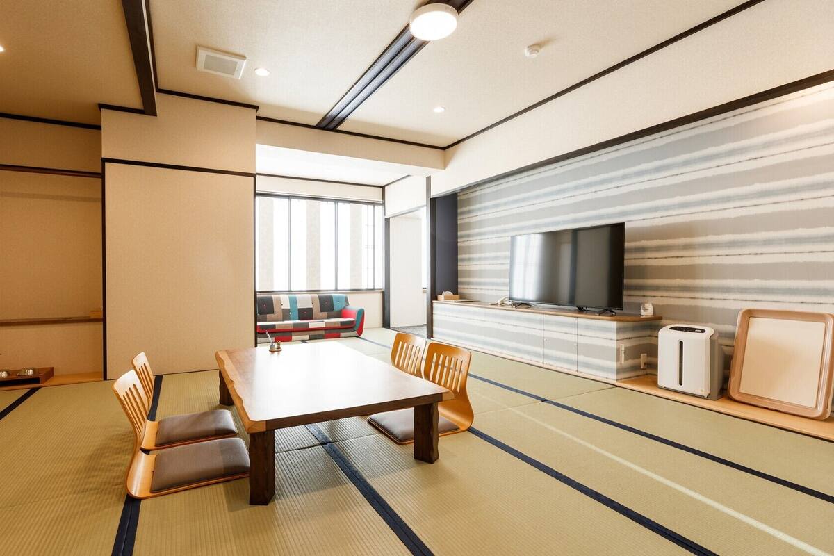 Una habitación de estilo japonés tradicional con suelo de tatami, una mesa baja con cojines, un sofá, un televisor y un refrigerador pequeño.
