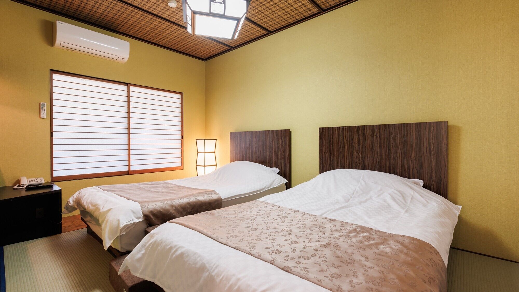 A hotel room with two beds, a bedside table with a phone, a wall-mounted air conditioner, and a window with blinds.