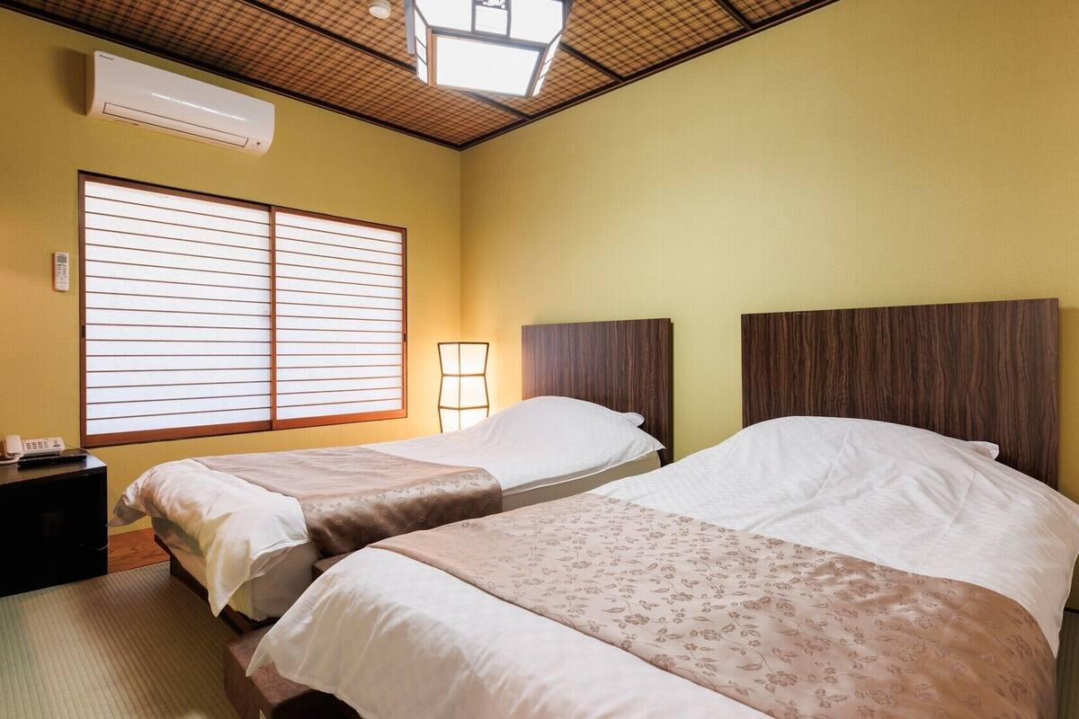 Habitación de hotel con dos camas, una mesita de noche con teléfono, aire acondicionado instalado en la pared y una ventana con persianas.