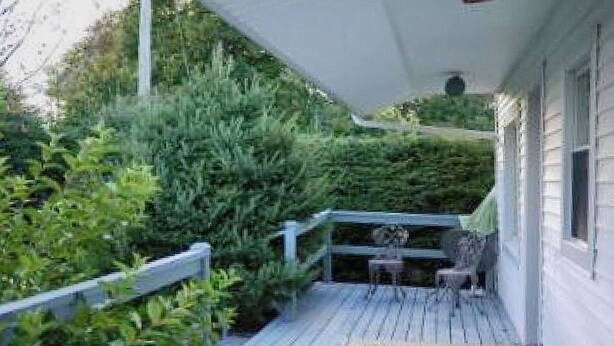 A wooden deck with a railing, surrounded by dense greenery. - Linden Homestead: King Bed Cottage on Century Farm Property (Stanley Bridge)
