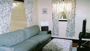 A living room with a sofa, a small table with a lamp, a television, and a window with curtains. - Linden Homestead: King Bed Cottage on Century Farm Property (Stanley Bridge)