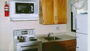 A compact kitchen with a white microwave, wooden cabinets, a stove, and a sink. - Linden Homestead: King Bed Cottage on Century Farm Property (Stanley Bridge)