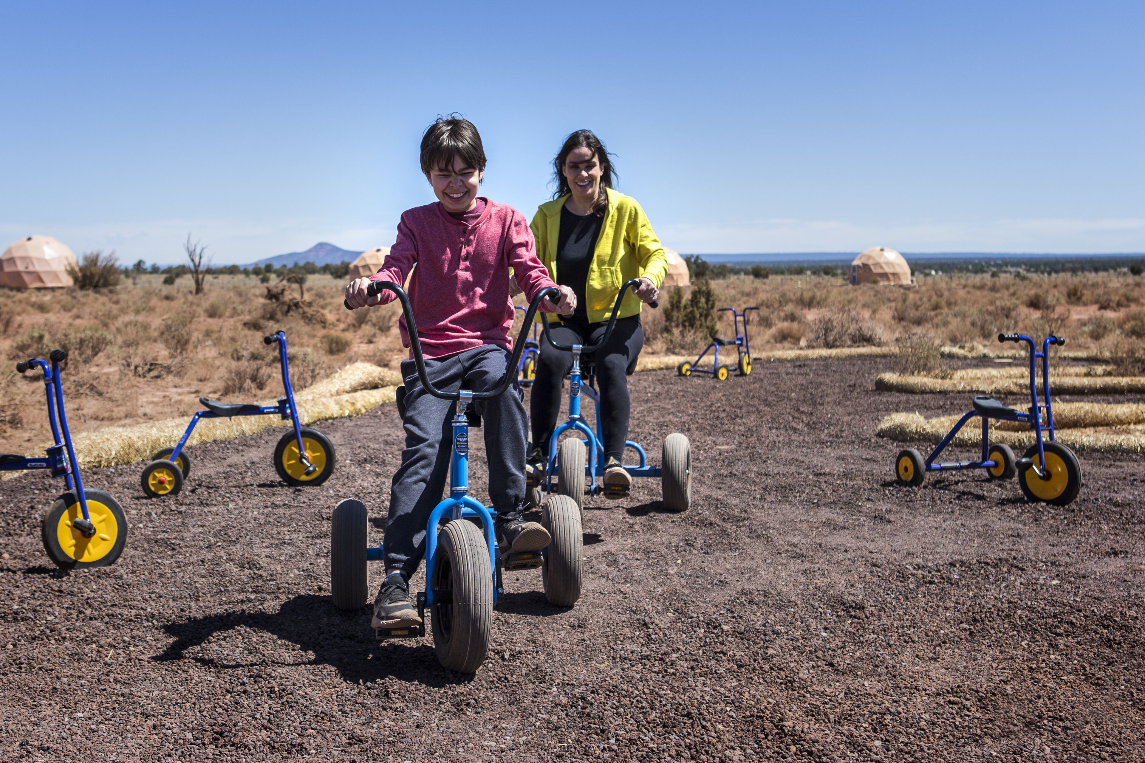 Dos personas paseando en triciclos por un campo con pacas de heno y triciclos azules al fondo.