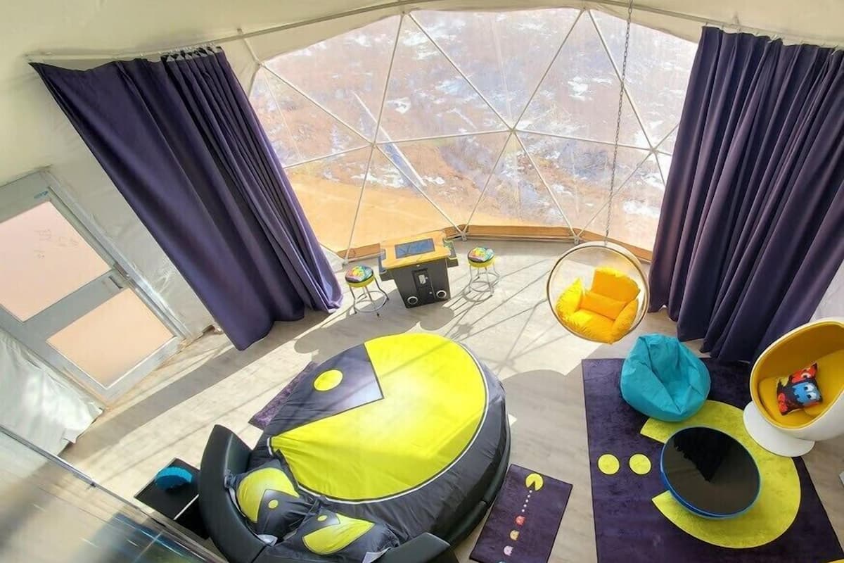 Une pièce spacieuse avec une grande fenêtre, un canapé rond jaune et noir, un fauteuil suspendu et une petite table avec une chaise.