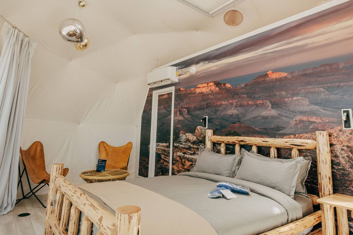 Une chambre avec un lit en bois, une lucarne, une grande fenêtre, une fresque représentant un canyon et une petite table.