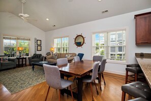 A spacious living room with a wooden dining table, grey chairs, a sofa, and a coffee table. There are large windows with blinds, a ceiling fan, and a wooden cabinet.