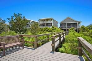 A wooden deck with a bench, surrounded by lush greenery and overlooking a beachfront property with multiple buildings. - Ale Of A Good Time: Modern Home Overlooking Canal (Oak Island)