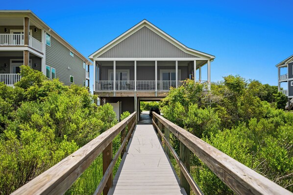 A wooden boardwalk leading to a building with a balcony, surrounded by dense greenery. - Ale Of A Good Time: Modern Home Overlooking Canal (Oak Island)