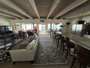 A spacious living room with a sofa, a coffee table, and a television. There is a kitchen area with a bar and chairs. The room has a large window and a ceiling with exposed beams.