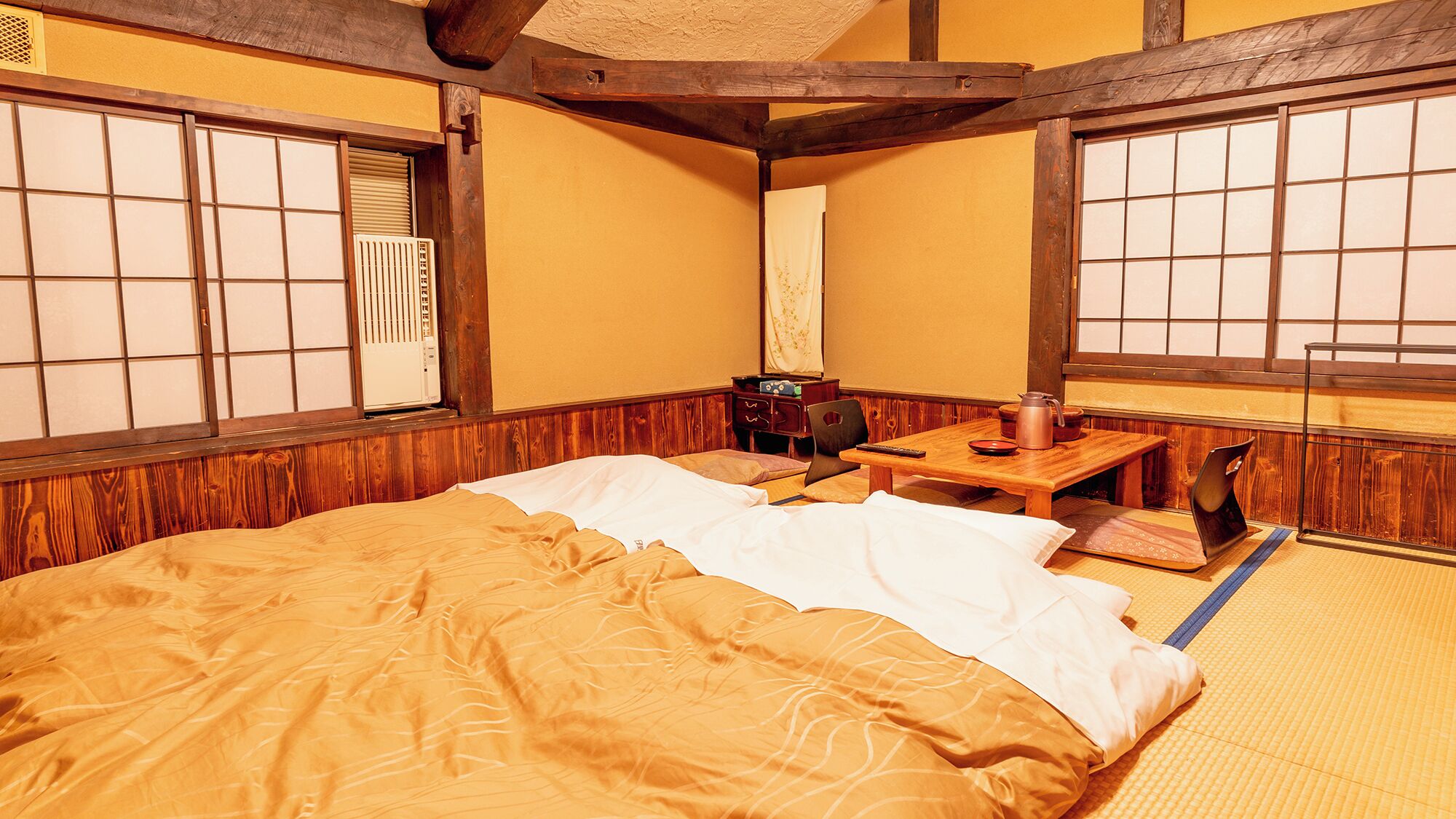 Ein traditioneller japanischer Raum mit Tatami-Boden, einem Bett mit weißer und brauner Decke, einem kleinen Holztisch mit Wasserkocher und einem Heizkörper.