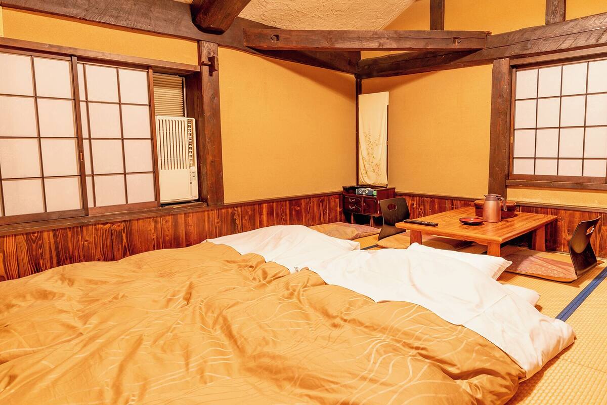Perinteinen japanilaistyylinen huone, jossa on tatamimatto, valko-ruskea peitto sängyllä, pieni puinen pöytä ja vedenkeitin sekä patteri.