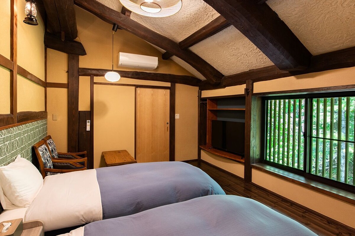 A room with two beds, a wooden ceiling, a window with a view of greenery, and a small table.