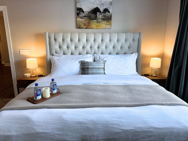A neatly made bed with white linens, a grey headboard, and a small bedside table with a lamp. There is a bottle of water and a glass on the bed.