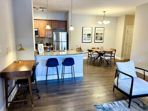A modern kitchen with stainless steel appliances, a dining area with a wooden table and chairs, and a small bar area with blue chairs.