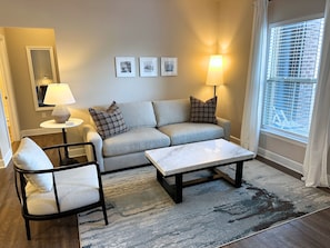 A living room with a sofa, two armchairs, a coffee table, a lamp, and a rug. There are three framed pictures on the wall.