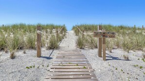 Ein Holzsteg führt zu einem Strandabschnitt mit einem Wegweiser und einem Seilzaun.