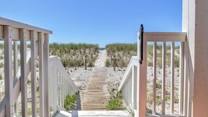 Ein hölzerner Weg führt zu einem Strand, gesäumt von weißen Geländern und umgeben von Sand und spärlicher Vegetation.