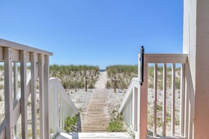 Un sentier en bois menant à une plage, bordé de rampes de garde-corps blanches et entouré de sable et d’une végétation clairsemée.