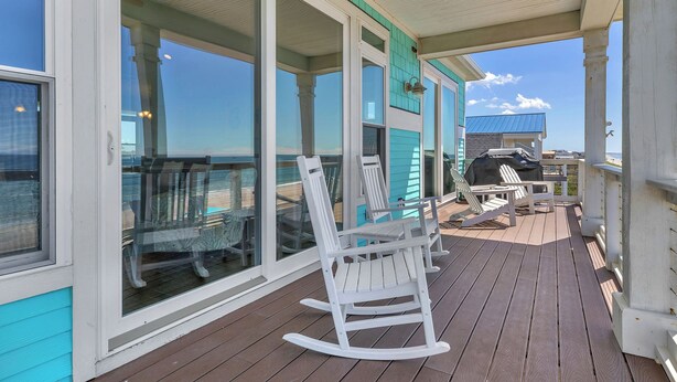 Eine überdachte Terrasse mit weißen Schaukelstühlen, einem Grill und einer Glastür, die zu einem Balkon führt.