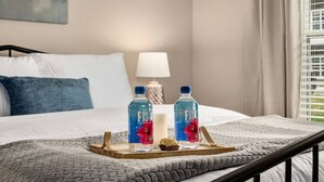 Un letto rifatto con cura, una coperta grigia, due bottiglie d'acqua Fiji e un piccolo muffin su un vassoio.