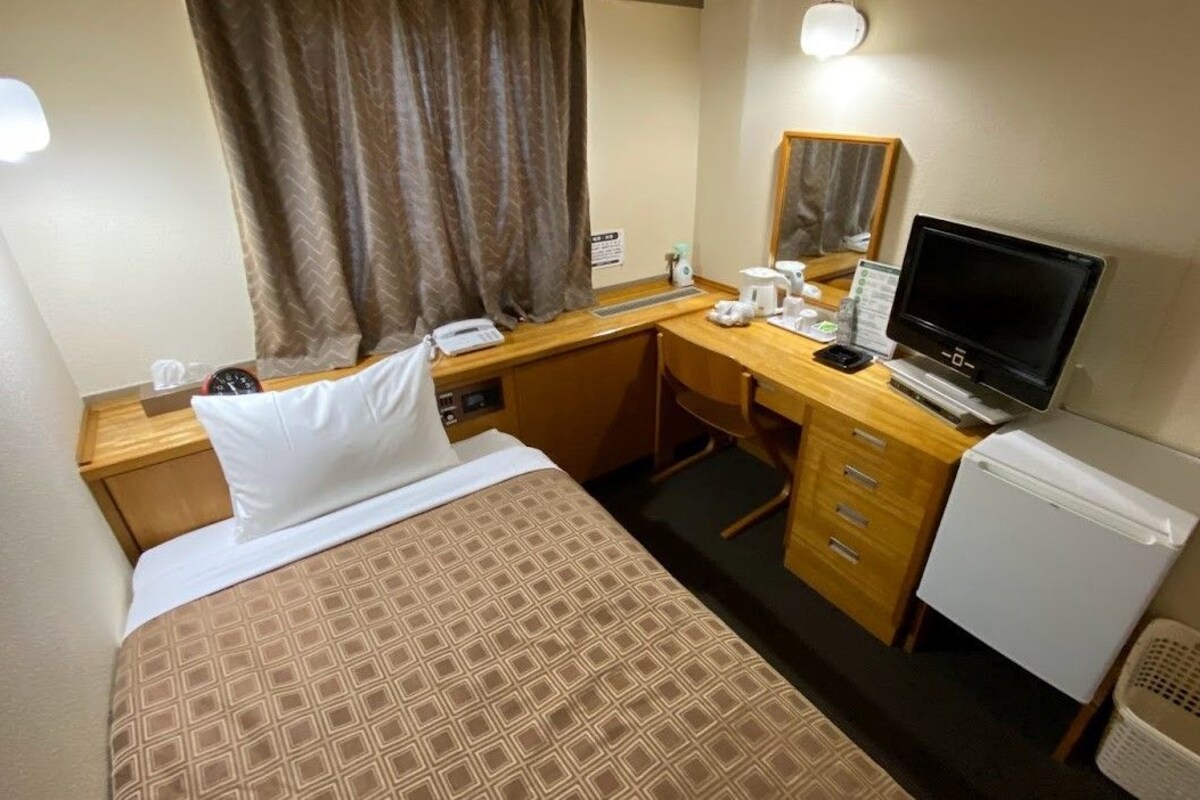 Habitación de hotel con una cama, un escritorio con computadora, un refrigerador y una ventana con cortinas.