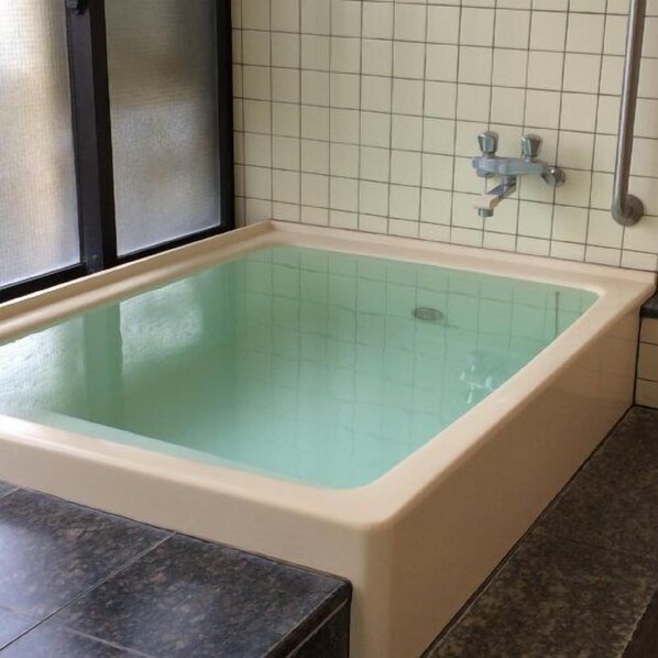 Una bañera rectangular con interior verde claro, un muro cubierto de azulejos con diseño de cuadrícula y un grifo metálico.