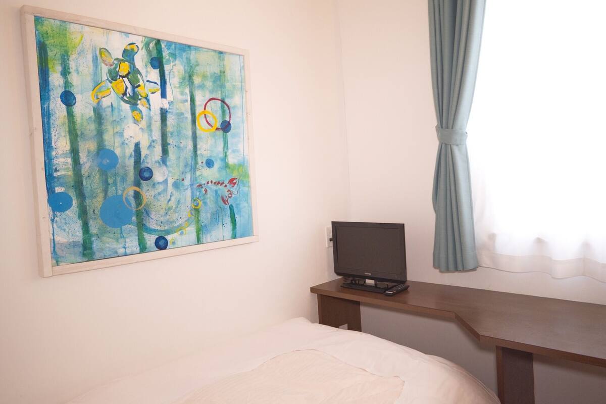 A bedroom with a bed, a desk with a computer, a painting on the wall, and a window with curtains.