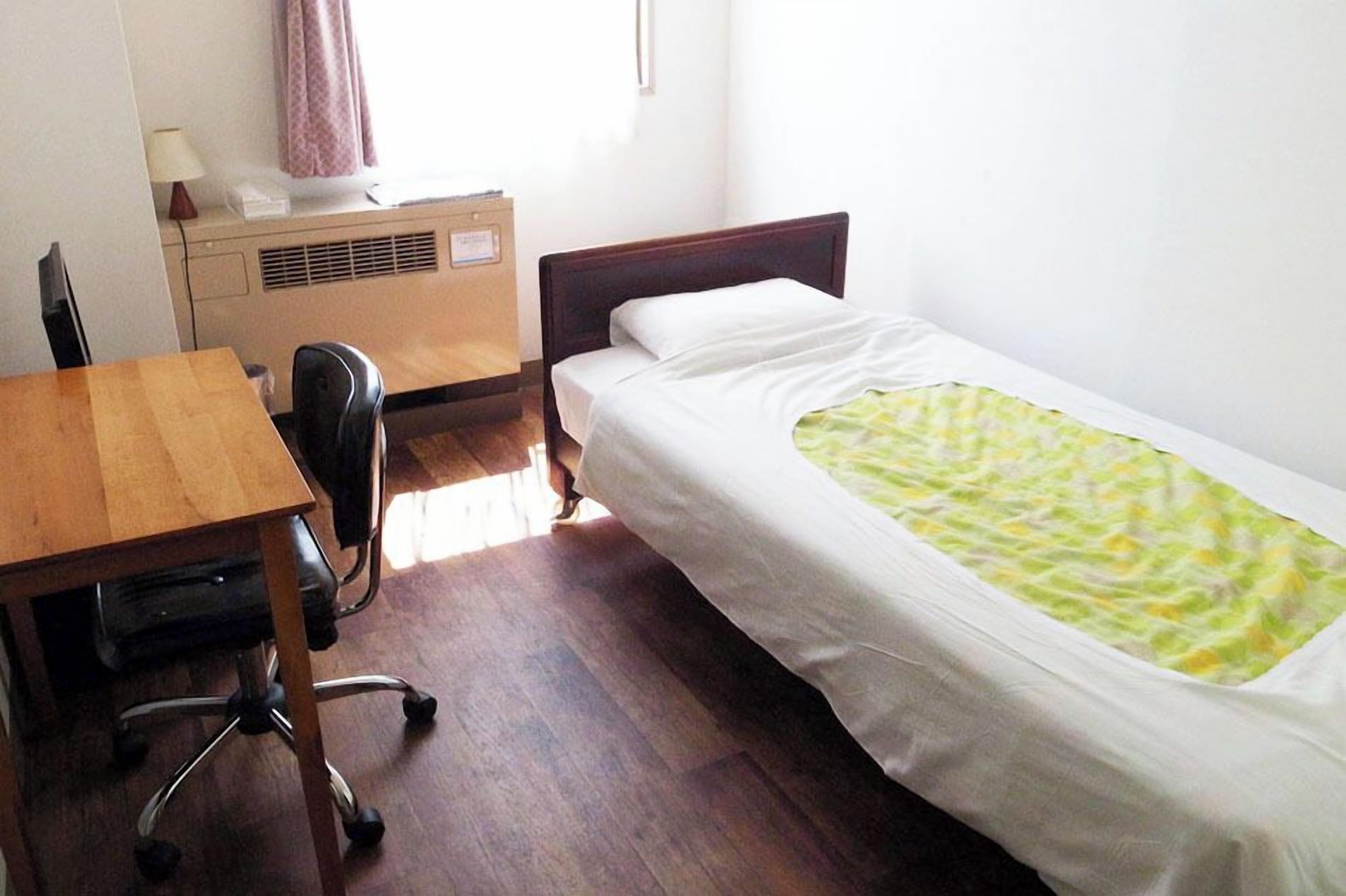 A single bed with a green patterned blanket, a wooden desk with a chair, and a small bedside table with a lamp.