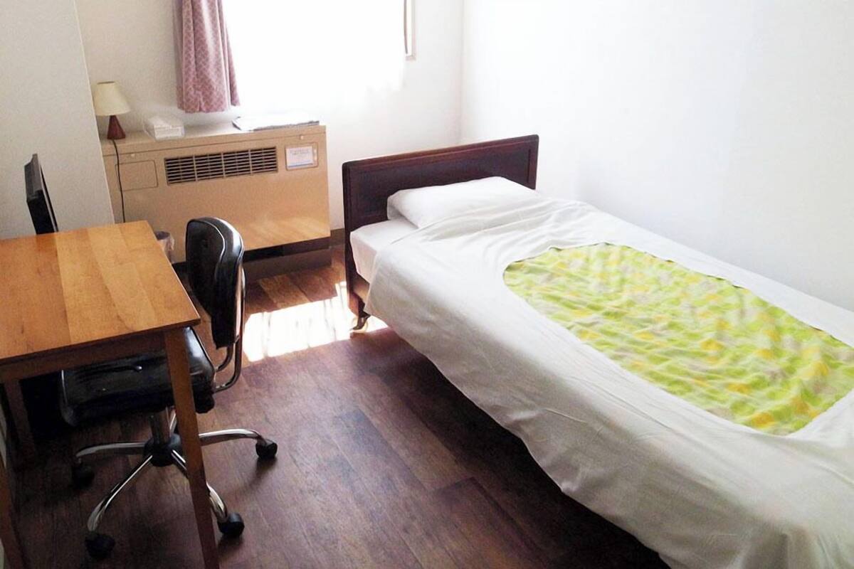 A single bed with a green patterned blanket, a wooden desk with a chair, and a small bedside table with a lamp.