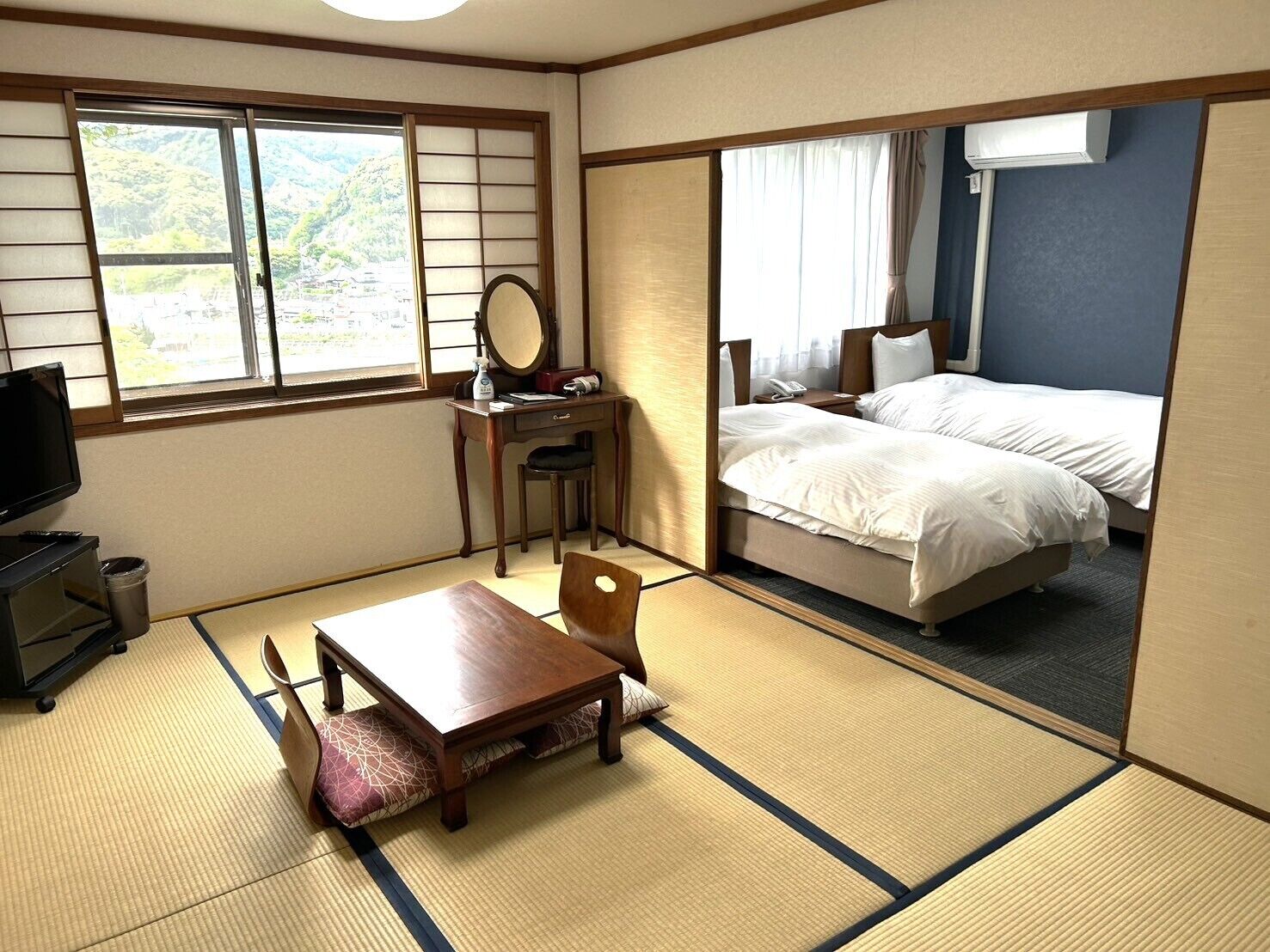 A traditional Japanese-style room with tatami flooring, a desk with a mirror, a bed with white bedding, and a television.