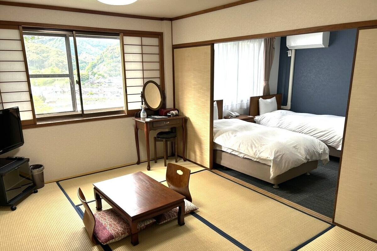 A traditional Japanese-style room with tatami flooring, a desk with a mirror, a bed with white bedding, and a television.