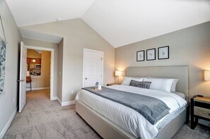 A bedroom with a large bed, a nightstand, a door leading to a bathroom, and a small kitchen area.
