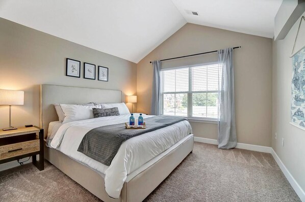 A bedroom with a large bed, bedside tables, a window with curtains, and framed artwork on the wall.