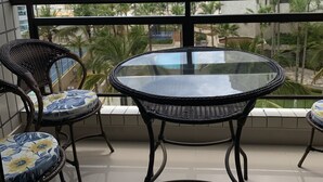 Un balcón con una mesa de vidrio y sillas de mimbre, con vista a una playa y palmeras.