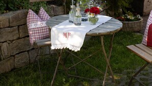 A wooden table set outdoors with a white tablecloth, surrounded by greenery and a stone wall.