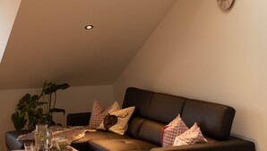 A cozy living room with a sloped ceiling, a dark leather sofa, a small wooden coffee table, and a potted plant.