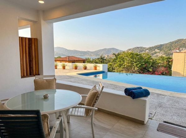 A balcony with a glass table and chairs, overlooking a swimming pool and a mountainous landscape.