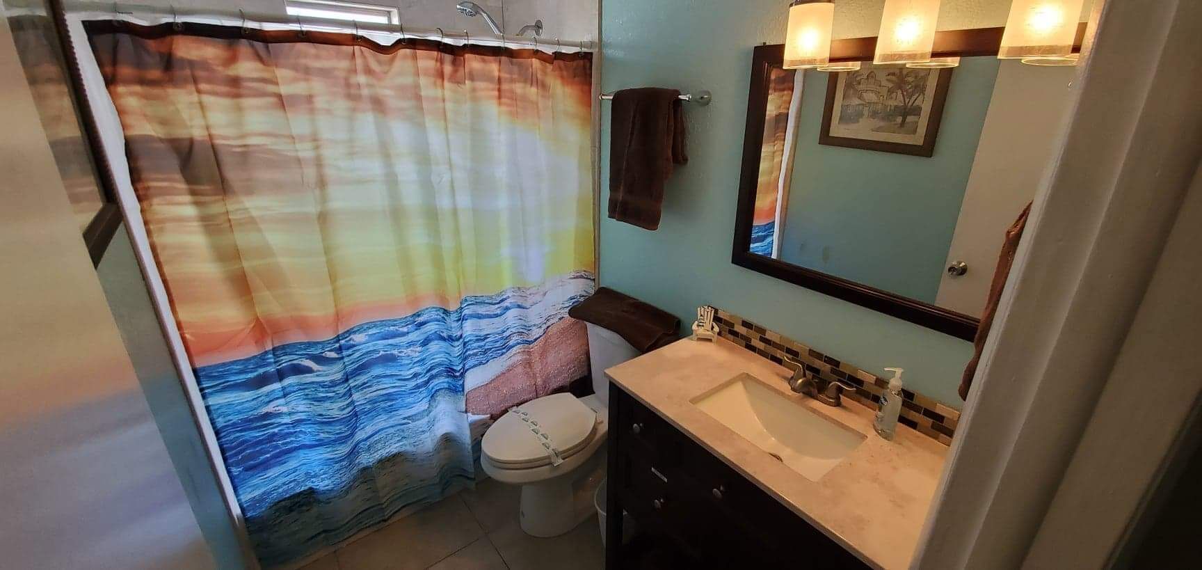 Une salle de bain avec un rideau de douche orné d’un motif de vagues, des toilettes, un lavabo avec un miroir au-dessus et une serviette accrochée au mur.