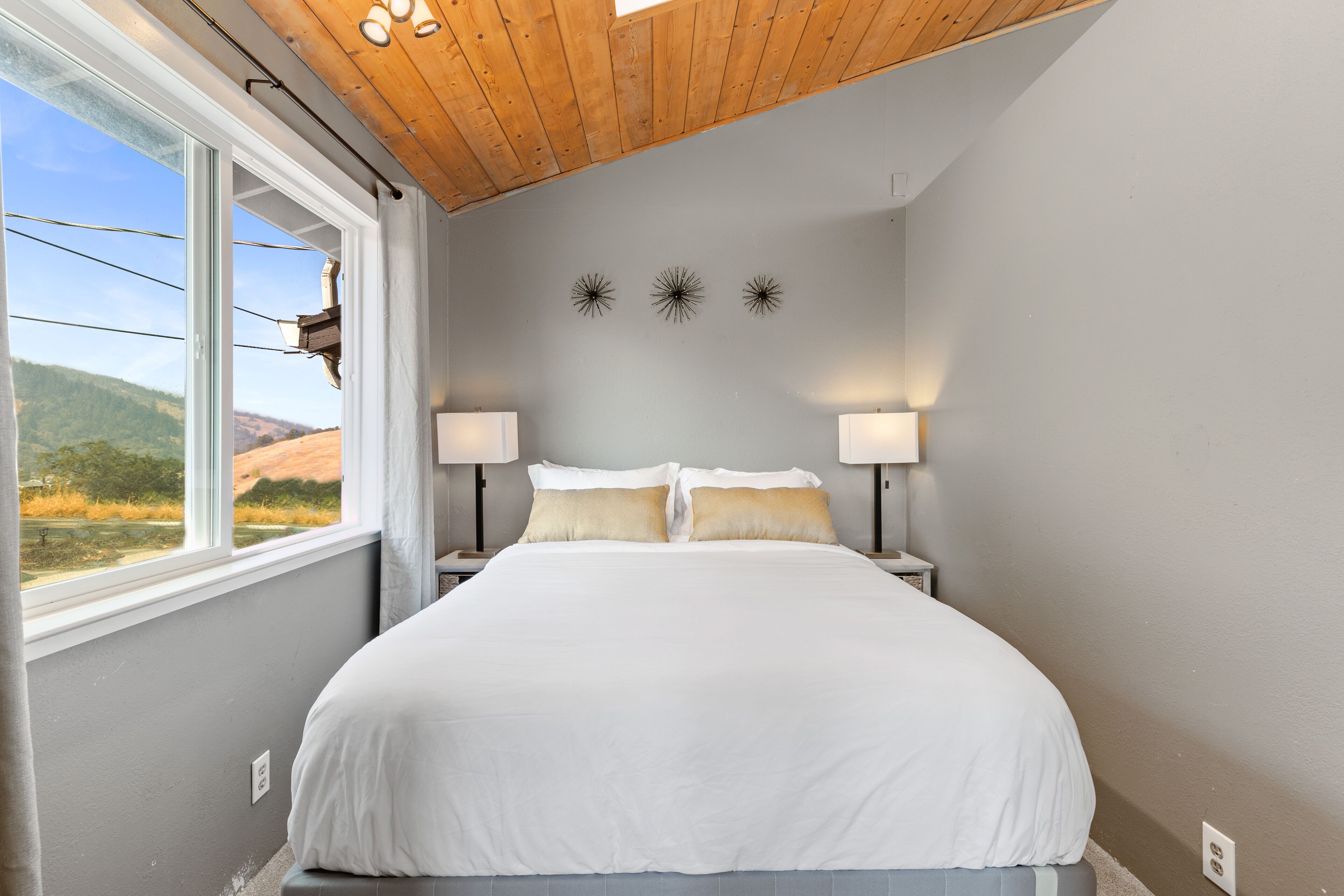 A bedroom with a large bed, two bedside lamps, a window with a view of the outdoors, and a wooden ceiling.