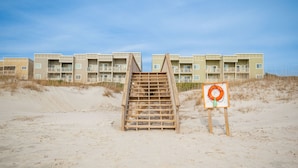 Eine Holztreppe führt zu einem Gebäude mit mehreren Balkonen, das direkt an einem Sandstrand mit einem Rettungsboje liegt.