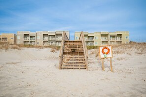 A wooden staircase leading to a building with multiple balconies, situated on a sandy beach with a lifebuoy on display.
