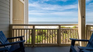Ein überdachter Balkon mit zwei Stühlen, der auf einen Strand mit Holzgeländer und Vegetation blickt.