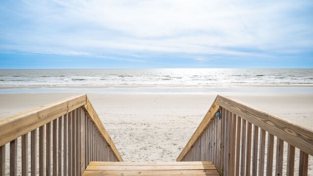 Ein Holzsteg führt zu einem Strand unter blauem Himmel.