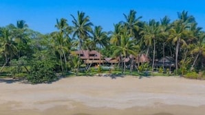 Een strand met helder turquoise water en palmbomen. In de verte zijn gebouwen met rieten daken te zien.
