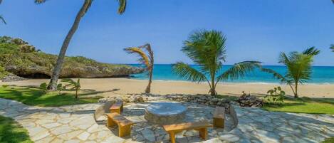 A stone patio with a fire pit and seating, surrounded by palm trees and a beach view.